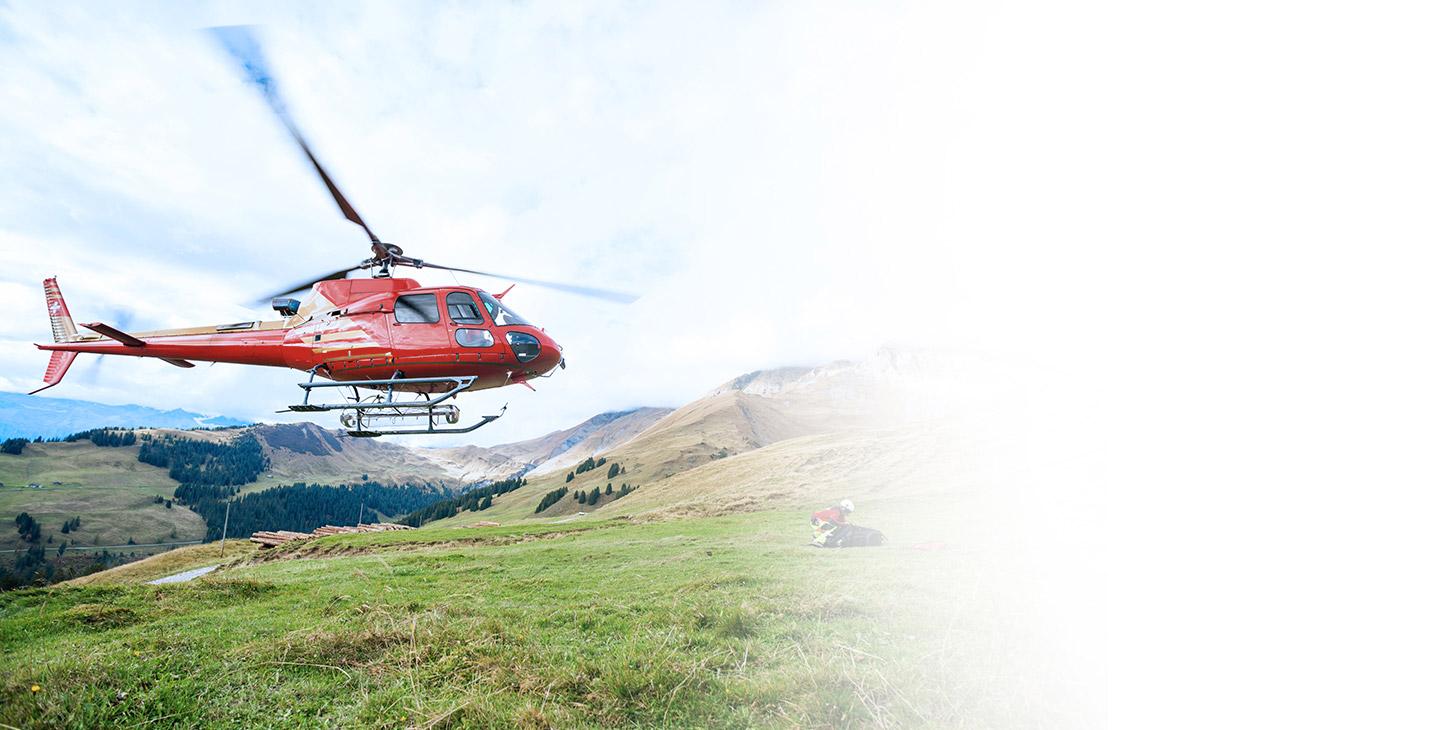 Red helicopter flying over a man kneeled down in the grass on the side of a mountain