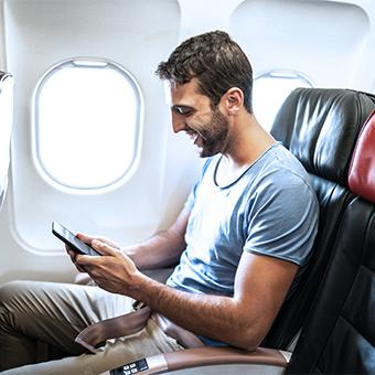 一名棕色头发的男子坐在飞机靠窗的位置上，用智能手机上网